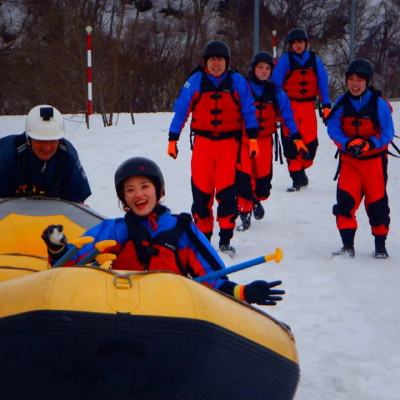 群馬県みなかみの冬のスノーラフティング体験で、雪の上でゴムボートを運ぶ参加者のグループ。, Snow rafting experience in Minakami, Japan, showing a cheerful group carrying a yellow raft across snowy terrain during a winter outdoor adventure, dressed in colorful cold-weather rafting gear.