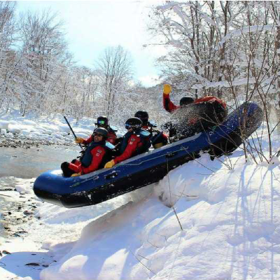 群馬県みなかみの冬のスノーラフティング体験。雪の斜面を川へ滑り降りるグループのアクティビティ。,Winter snow rafting adventure in Minakami, Japan, showing a group of rafters sliding down a snowy riverbank into an icy stream, enjoying an extreme outdoor winter activity in full safety gear.