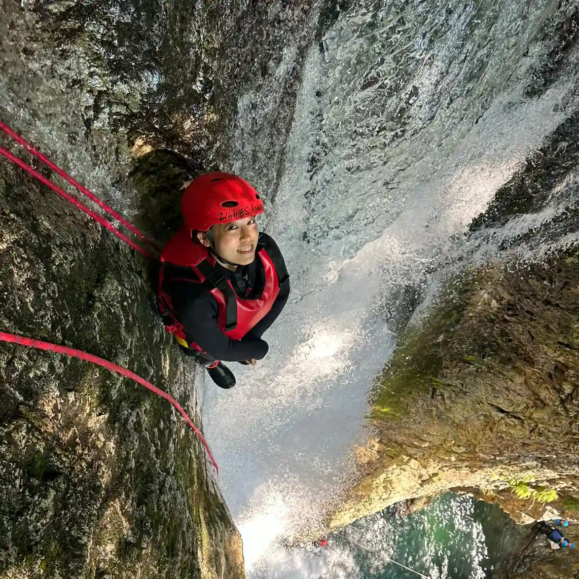みなかみフォックスキャニオニング冒険者が天然の渓谷プールの上を赤い安全ロープで滝を下降する空撮, Aerial view of minakami fox canyoning adventurer descending waterfall with red safety ropes over natural canyon pool.
